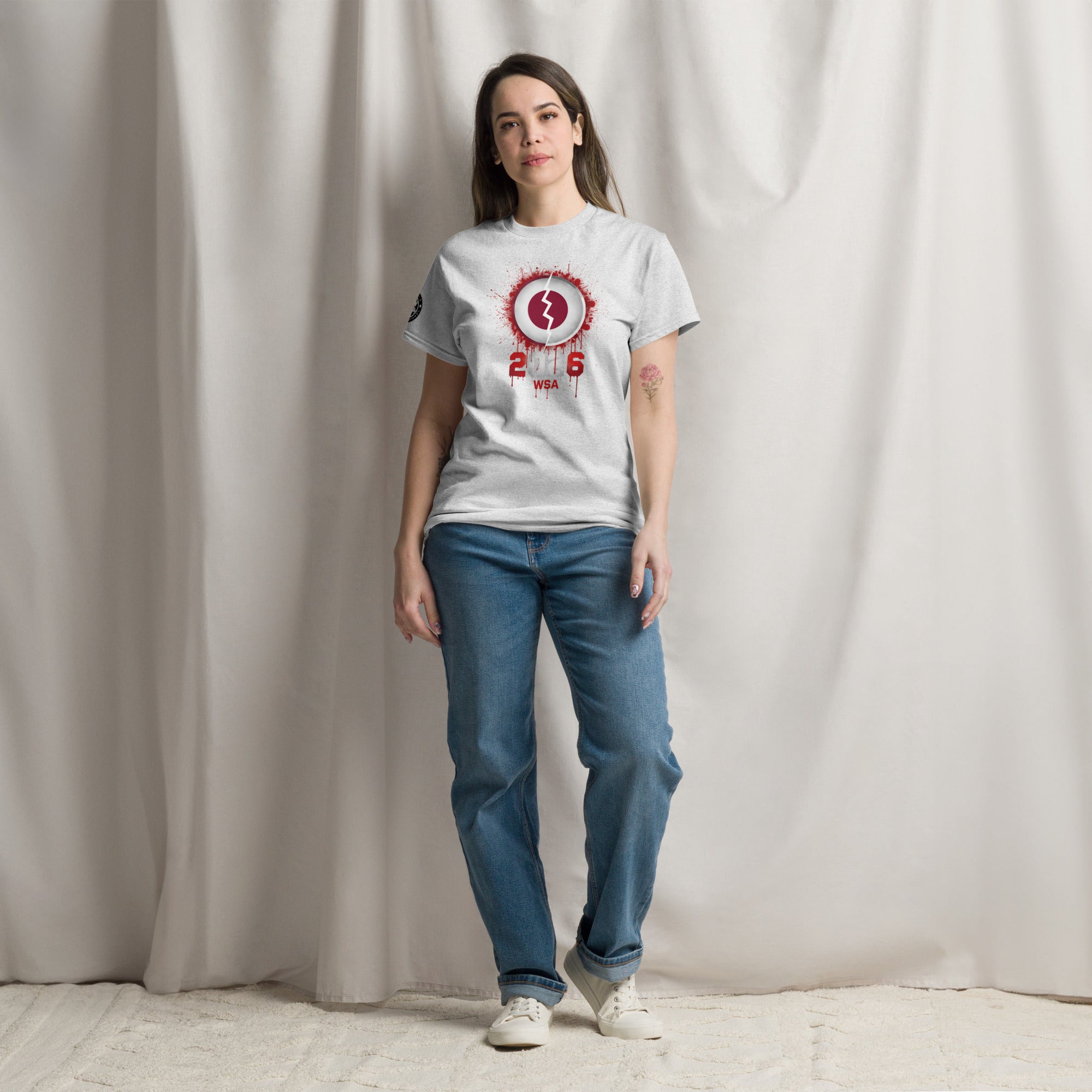 Person wearing a white t-shirt with a red and black design and blue jeans against a plain background