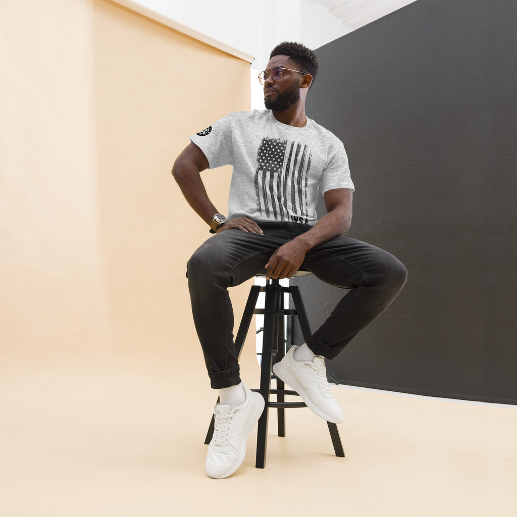 Man sitting on a stool wearing a white t-shirt with an American flag design, black pants, and white sneakers.
