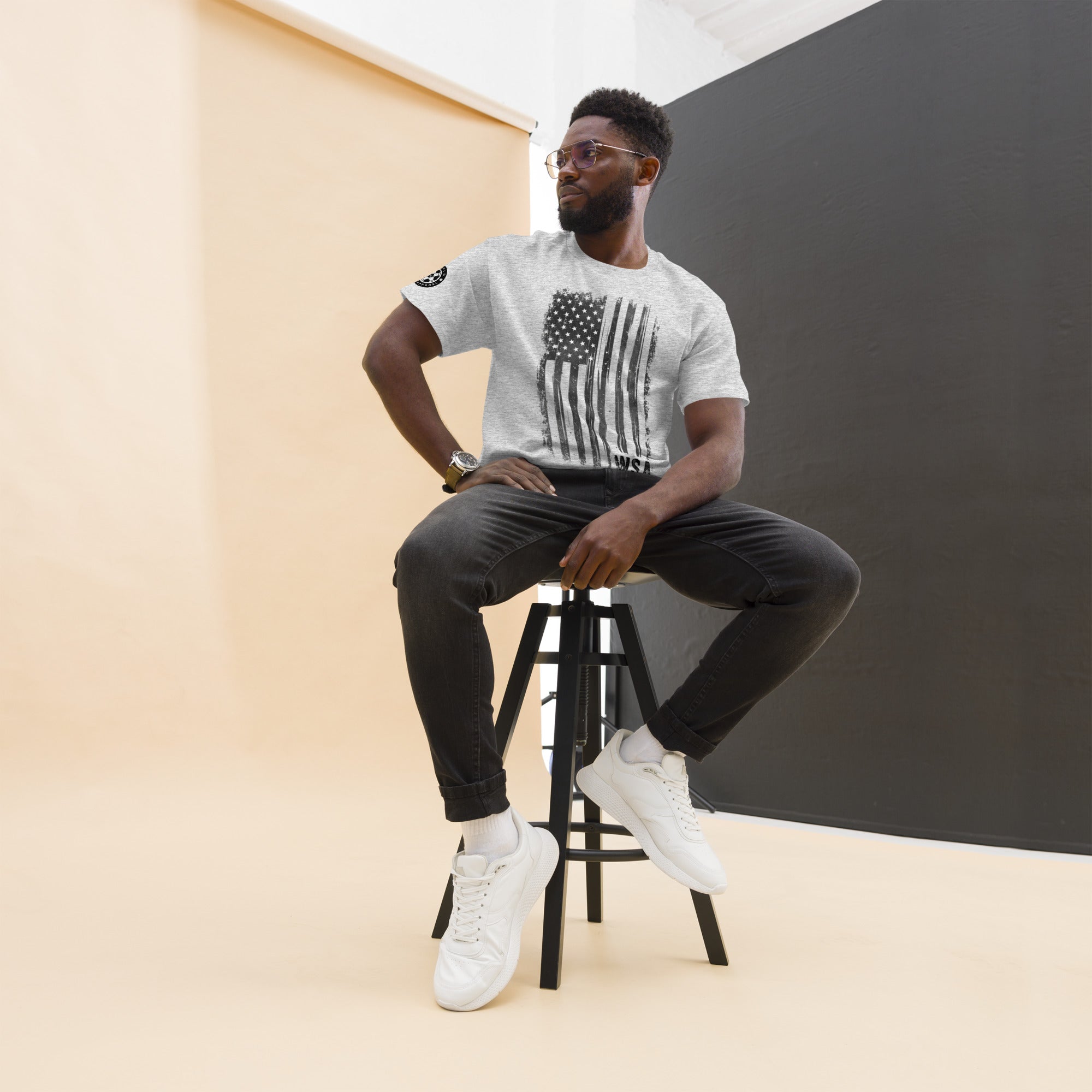 Man sitting on a stool wearing a white t-shirt with an American flag design, black pants, and white sneakers.