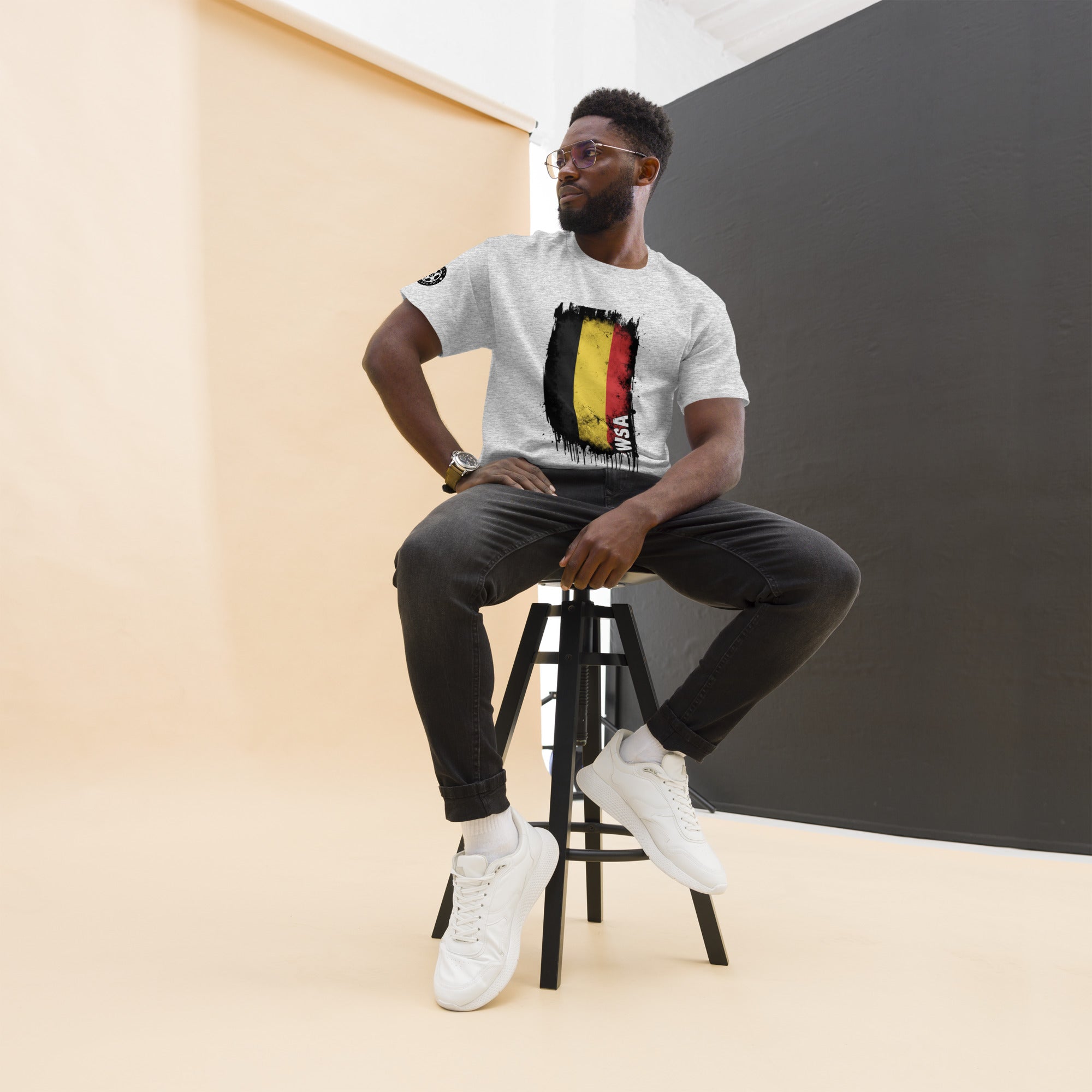 Man sitting on a stool wearing a white t-shirt with a graphic design, black pants, and white sneakers.
