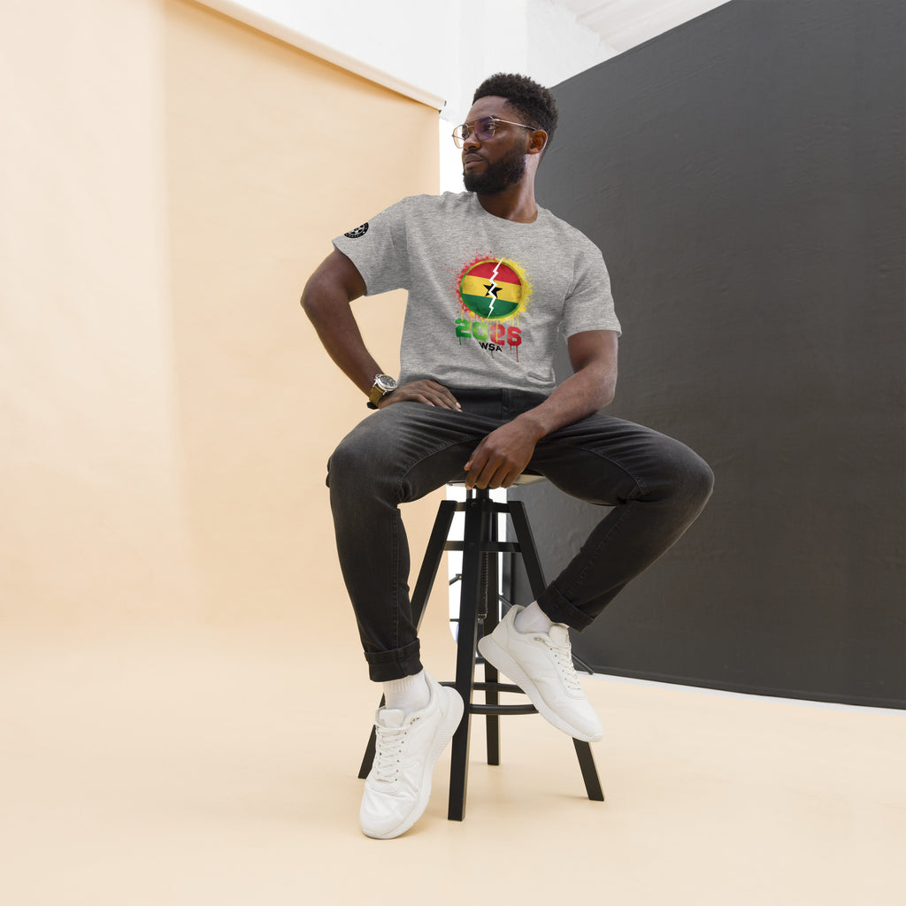 Man sitting on a stool wearing a gray t-shirt with a colorful design, black pants, and white sneakers.