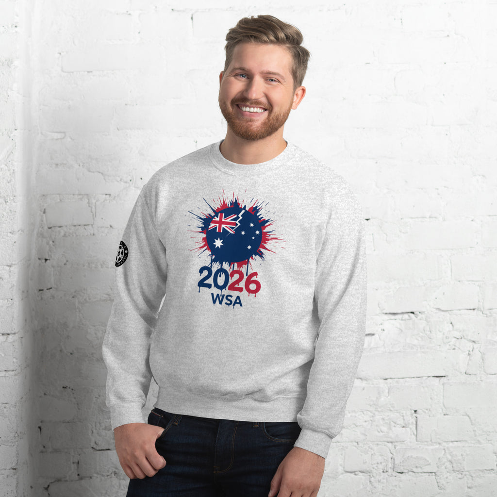 Man wearing a gray sweatshirt with a design featuring the number 2026 and text, standing against a white brick wall.