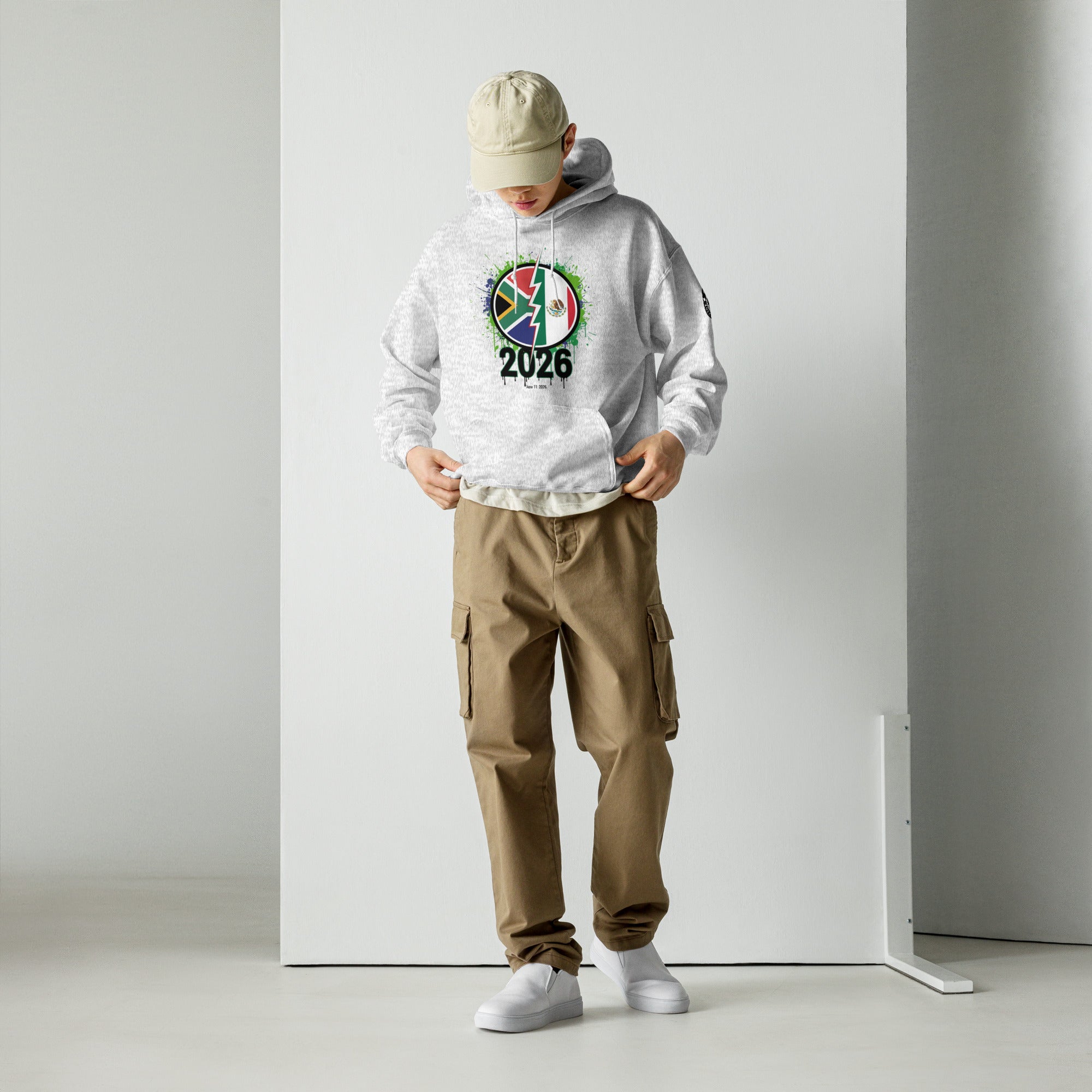 Person wearing a white hoodie with a colorful design and '2026' text, brown cargo pants, and a beige cap in a minimalistic setting.