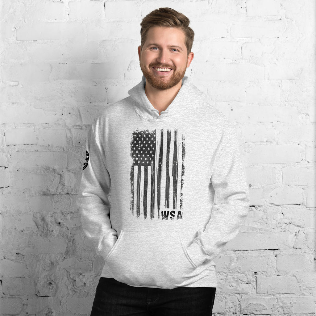 Man wearing a white hoodie with an American flag design against a white brick wall.
