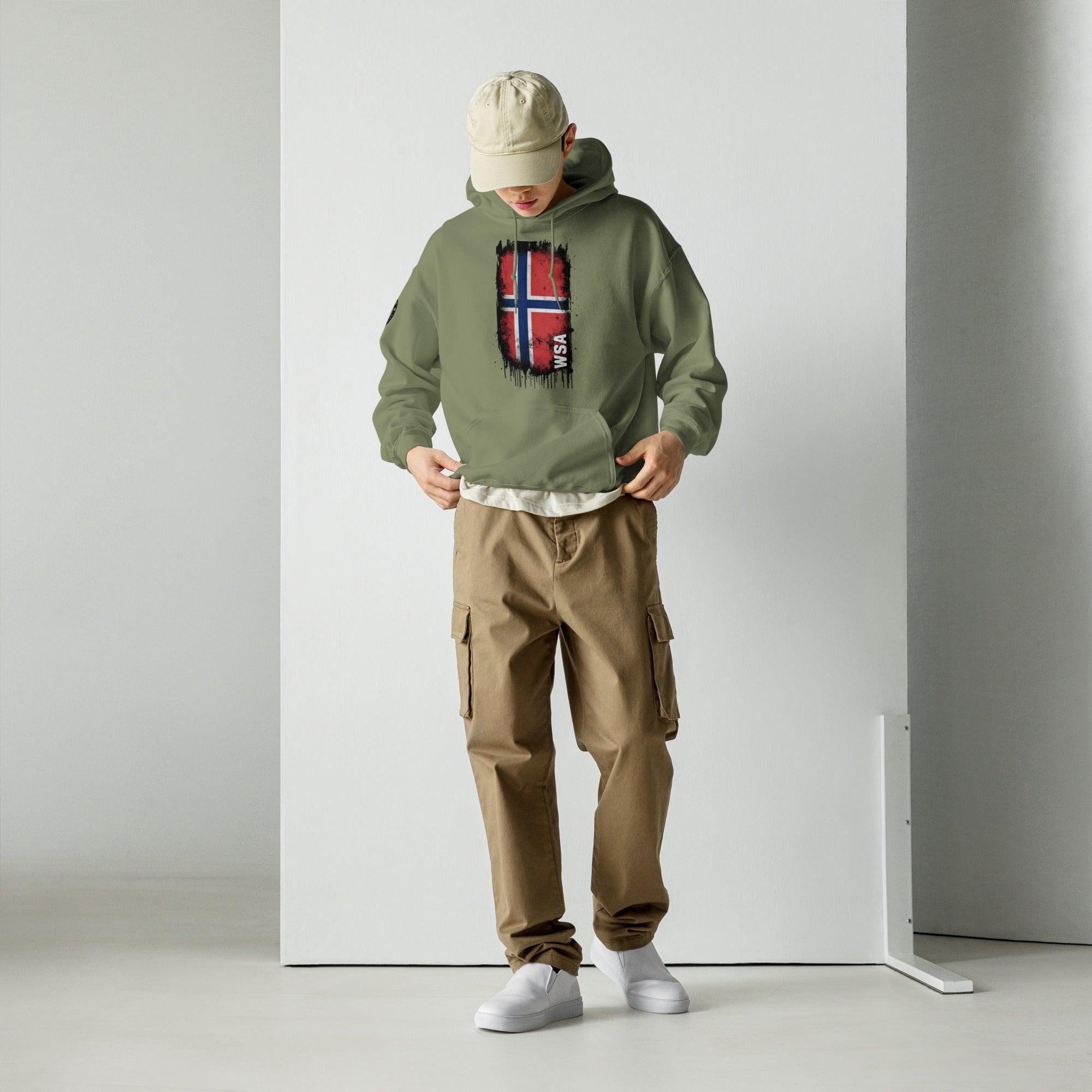 Person wearing a green hoodie with a red and blue design, beige cap, brown cargo pants, and white shoes against a plain background.