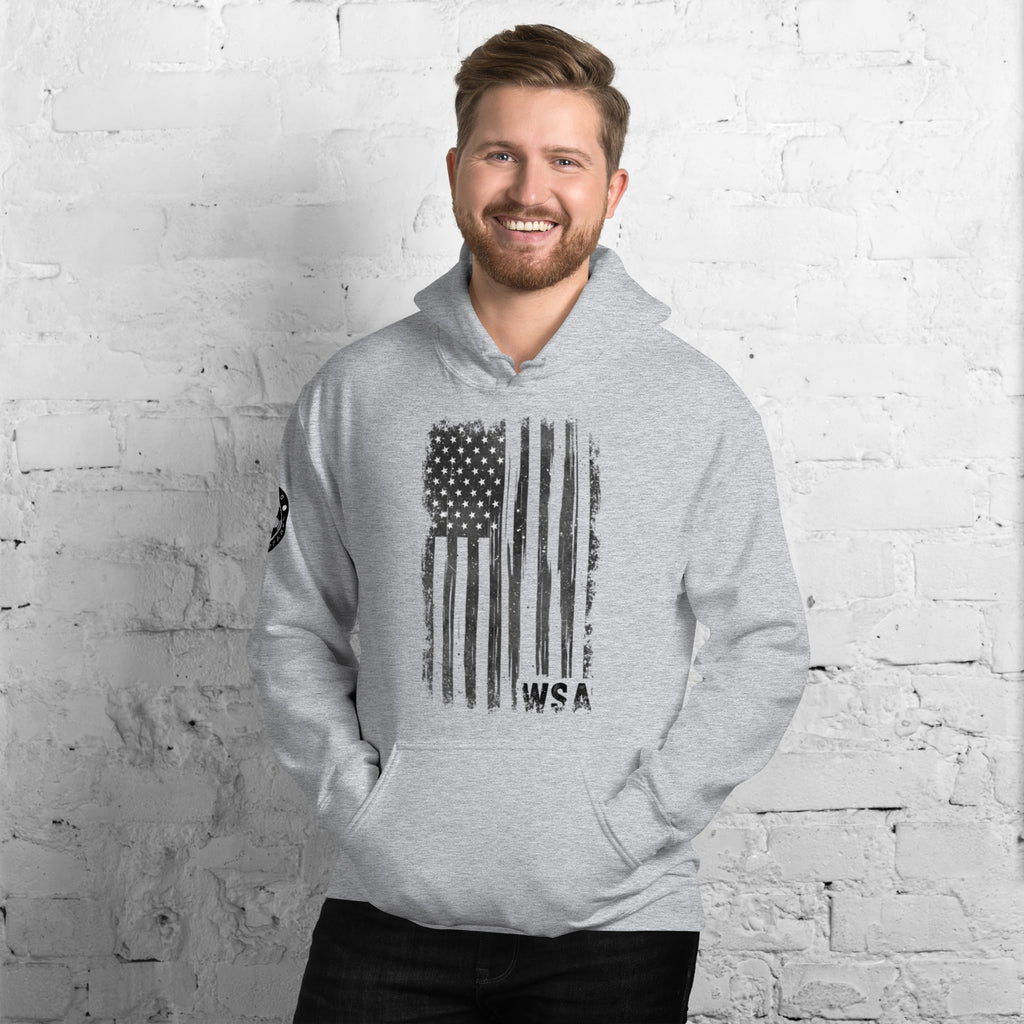 Man wearing a gray hoodie with an American flag design against a white brick wall.