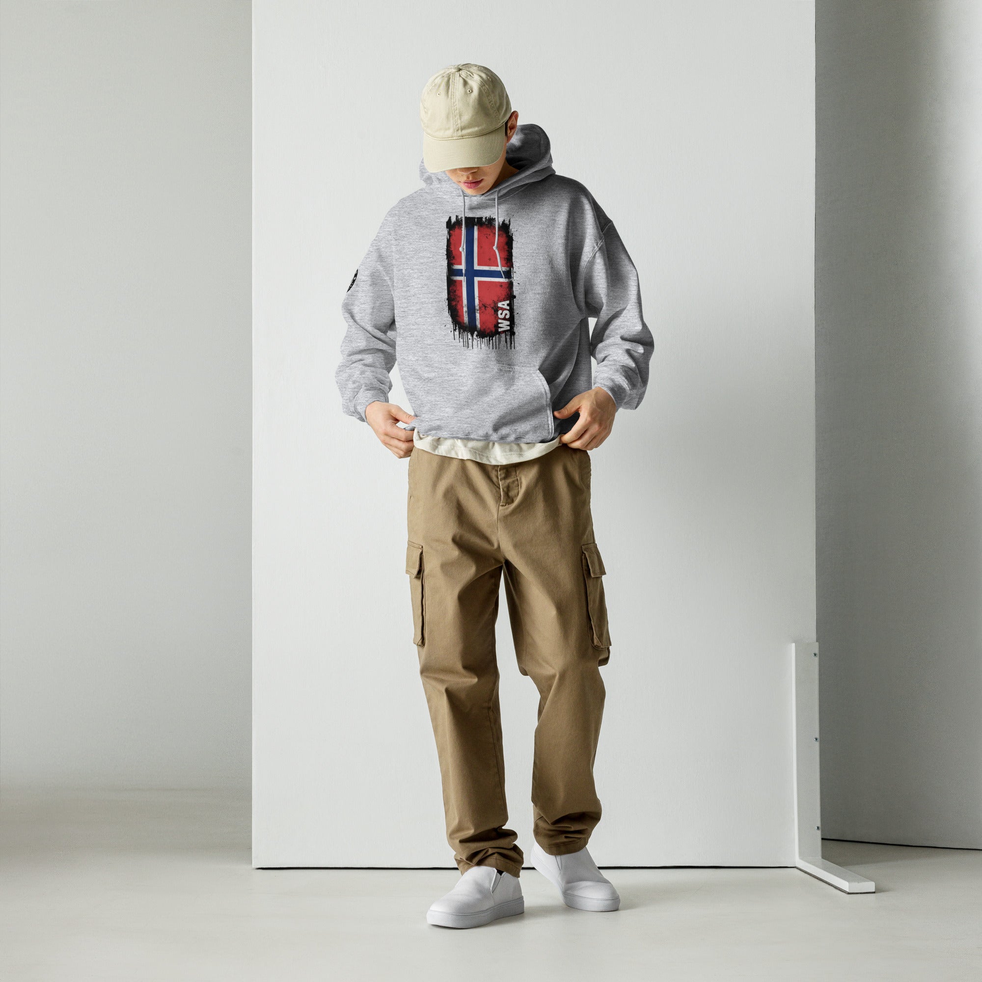 Person wearing a gray hoodie with a flag design, beige cap, and brown cargo pants against a white background.