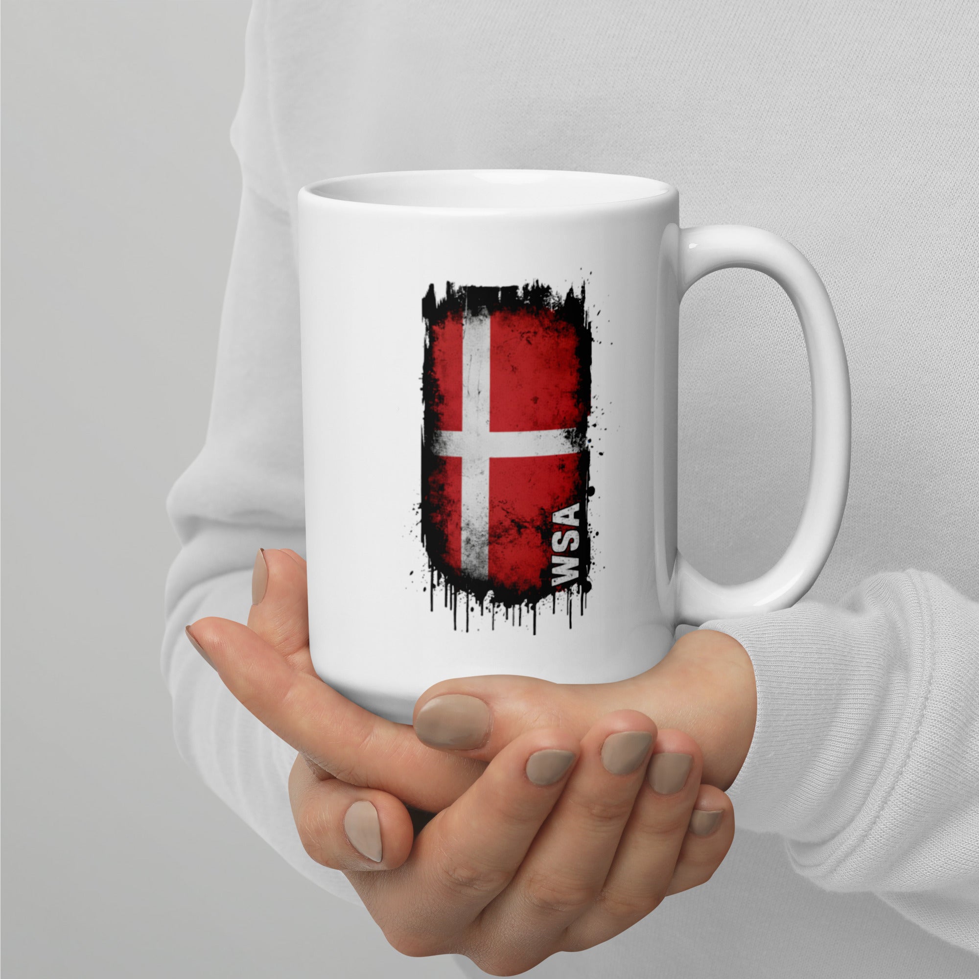 White mug with a flag design held by a person against a gray background