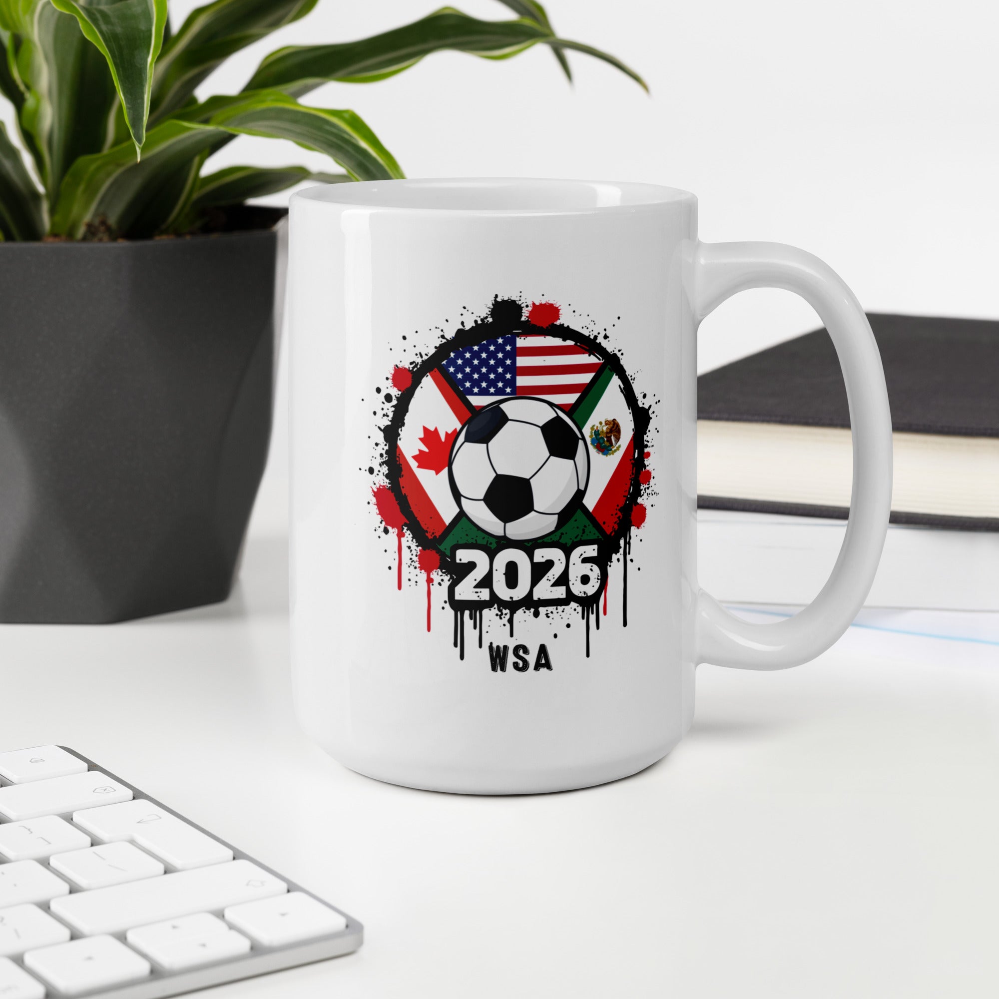 White mug with soccer ball design featuring flags and '2026 WSA' text on a desk with a plant and books in the background.