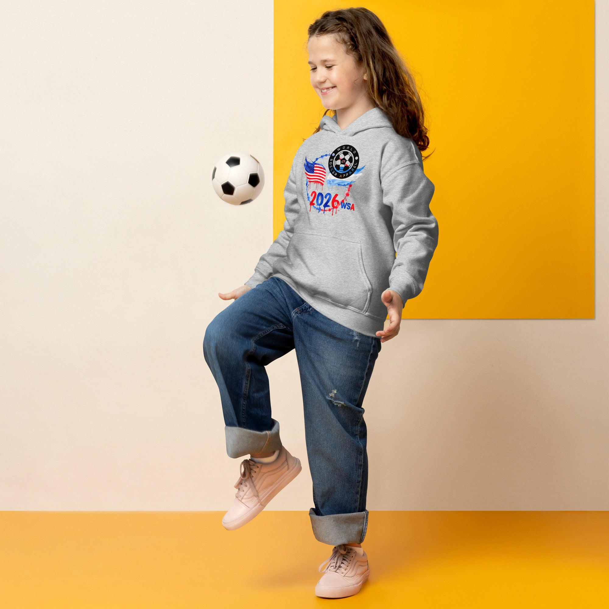Child wearing a gray hoodie with a soccer design, kicking a soccer ball against a yellow and beige background